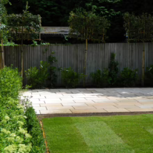 Landscaped backyard with a stone patio, manicured green lawn, and a wooden fence backdrop. Topiary trees and lush bushes add texture and depth to the garden design.
